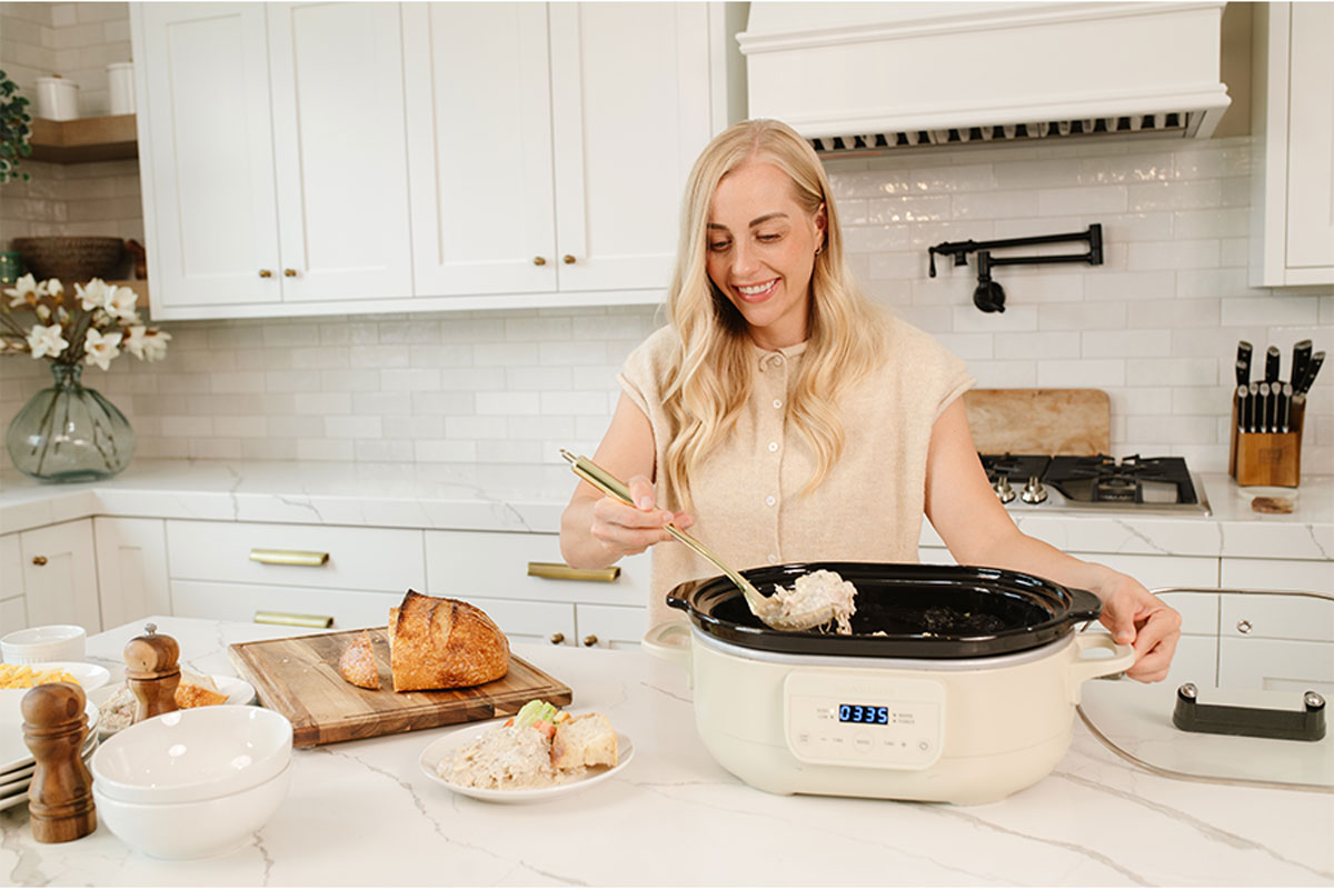 Woman scooping shredded chicken mixture out of slow cooker onto a plate in a white kitchen.