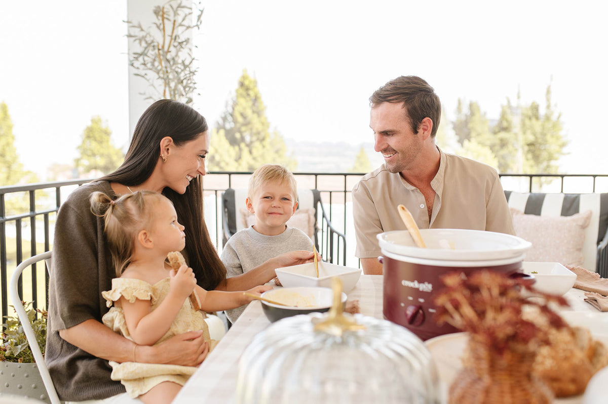 A family of four shares a meal on a sunny outdoor patio. A mother in a dark brown top holds a young toddler in a cream dress, while a father in a beige shirt sits next to them.