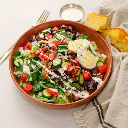 Salad with bbq pulled pork, vegetables, and hard boiled eggs in a brown bowl on a white surface.