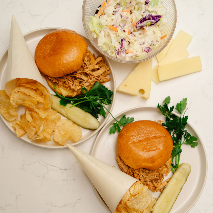 Two plates of sandwiches with sides including coleslaw, chips, and pickles on a light surface.