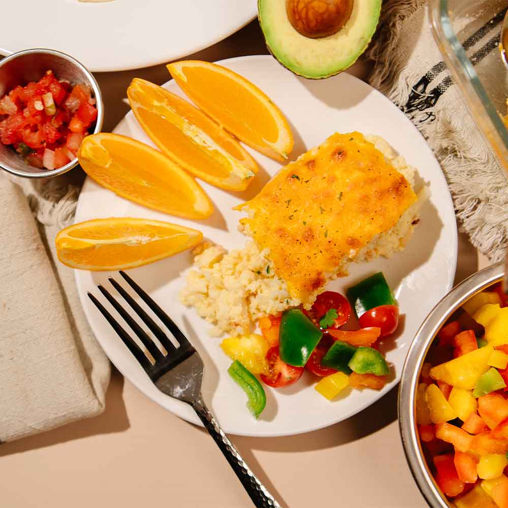 Plate of Beehive Meals Breakfast Casserole with orange slices, bell peppers, tomatoes and avocado with a fork on a beige background.