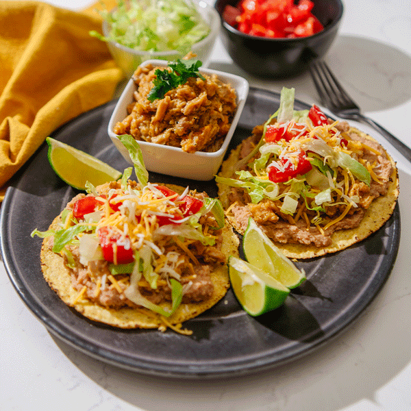 Two plated tostadas topped with lettuce, tomatoes and cheese with lime wedges on the side all on a black plate.