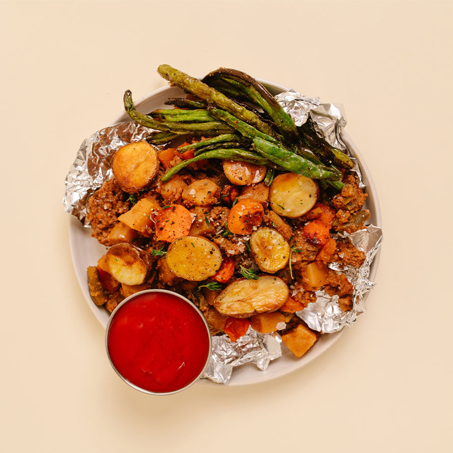 A top-down view of a plated meal on a light beige background. The food consists of roasted potatoes, carrots, green beans, and a browned ground beef, served on aluminum foil with a small metal cup of red dipping sauce.