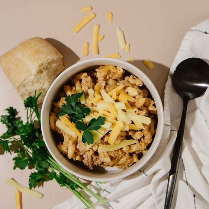 Bowl of Beehive Meals Cheeseburger Mac topped with shredded cheddar cheese with bread and parsley on a beige background