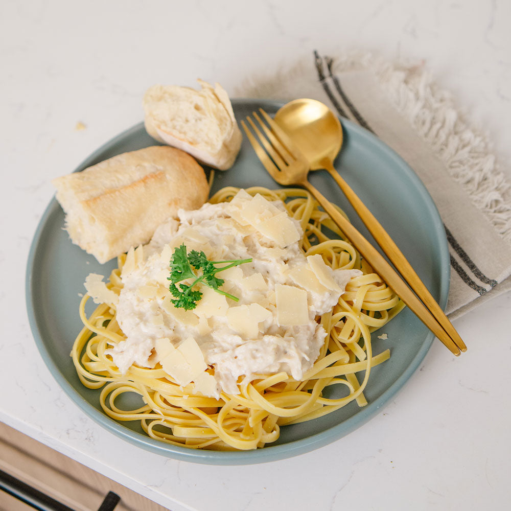 Plate of Beehive Meals Chicken Alfredo over fehttuchini noodles with bread and a gold fork and spoon on a white marble countertop. 