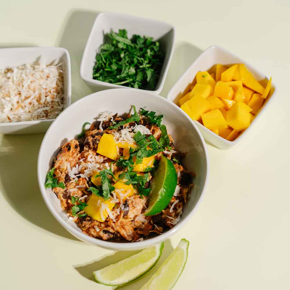Bowl of Beehive Meals Chicken Burrito Bowls in a white bowl topped with a lime slice, diced mangos, shredded coconut and cilantro with three little white bowls filled with shredded coconut, cilantro and chunks of mango on a light green background.