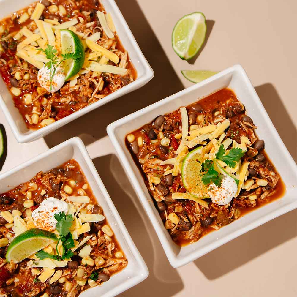 Three bowls of Beehive Meals Chicken Enchilada Soup with toppings on a beige background