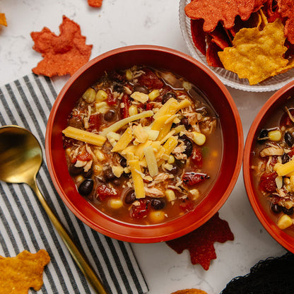 Red bowl of chicken tortilla soup with cheese, surrounded by tortilla chips on a white surface.