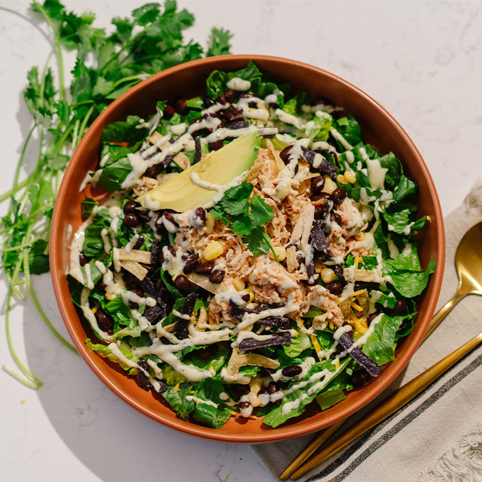 Salad with chicken, greens, avocado, and dressing in a brown bowl on a light surface.