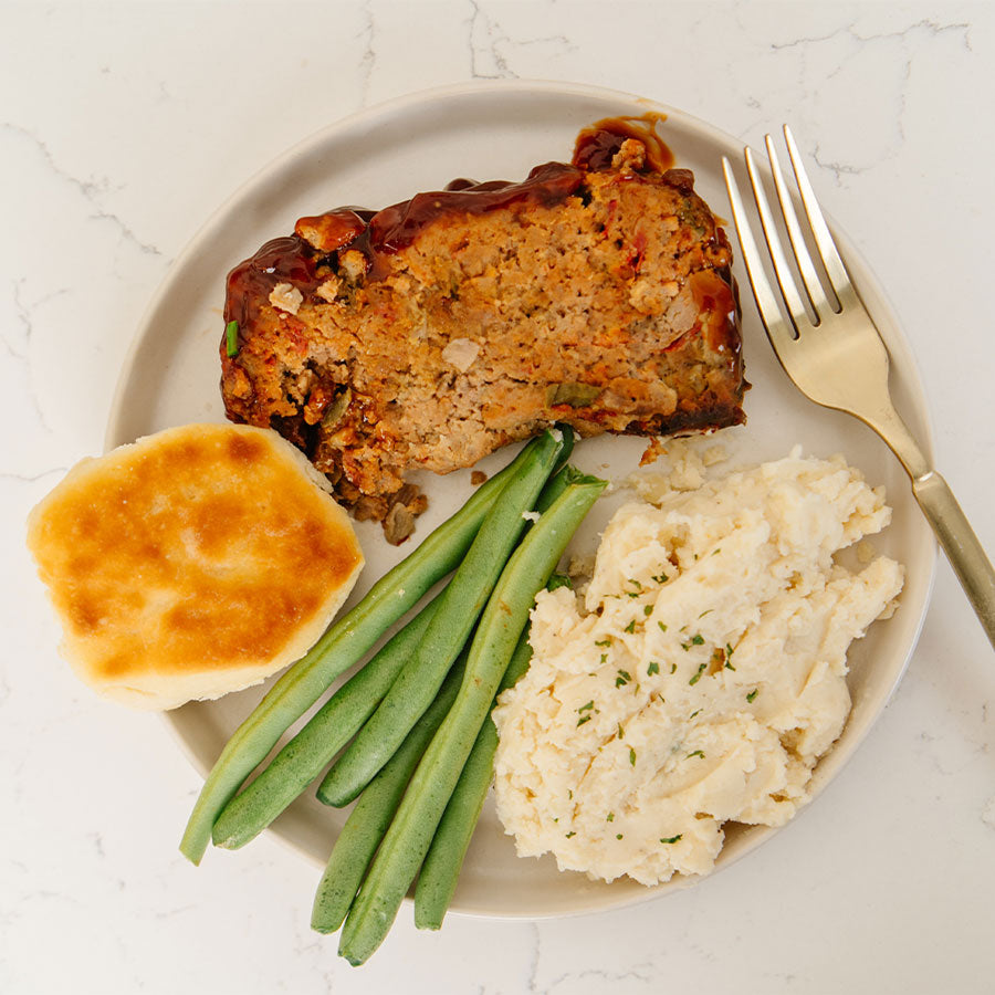 Plate of Beehive Meals Classic Meatloaf, green beans, mashed potatoes, and a biscuit on a white plate with a fork.
