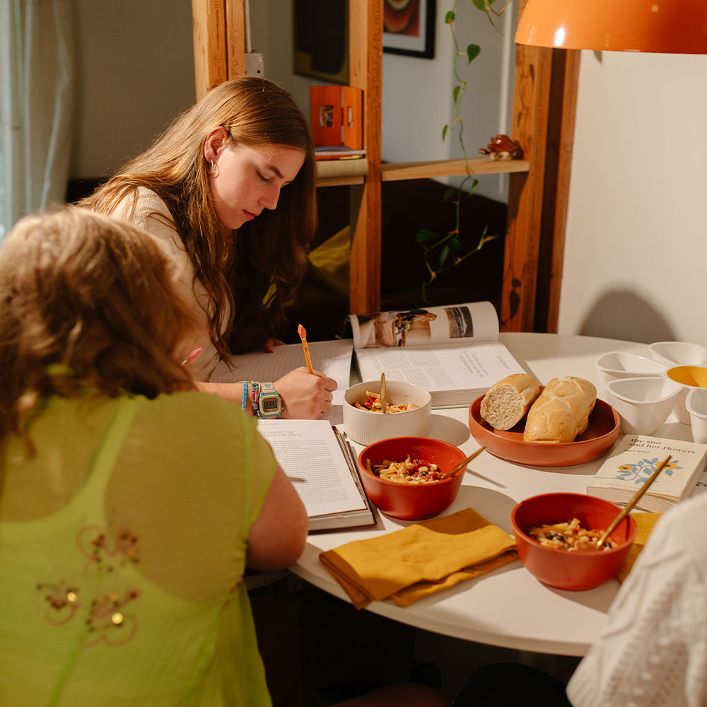 Two people sitting at a table with food and notebooks, engaged in a discussion.
