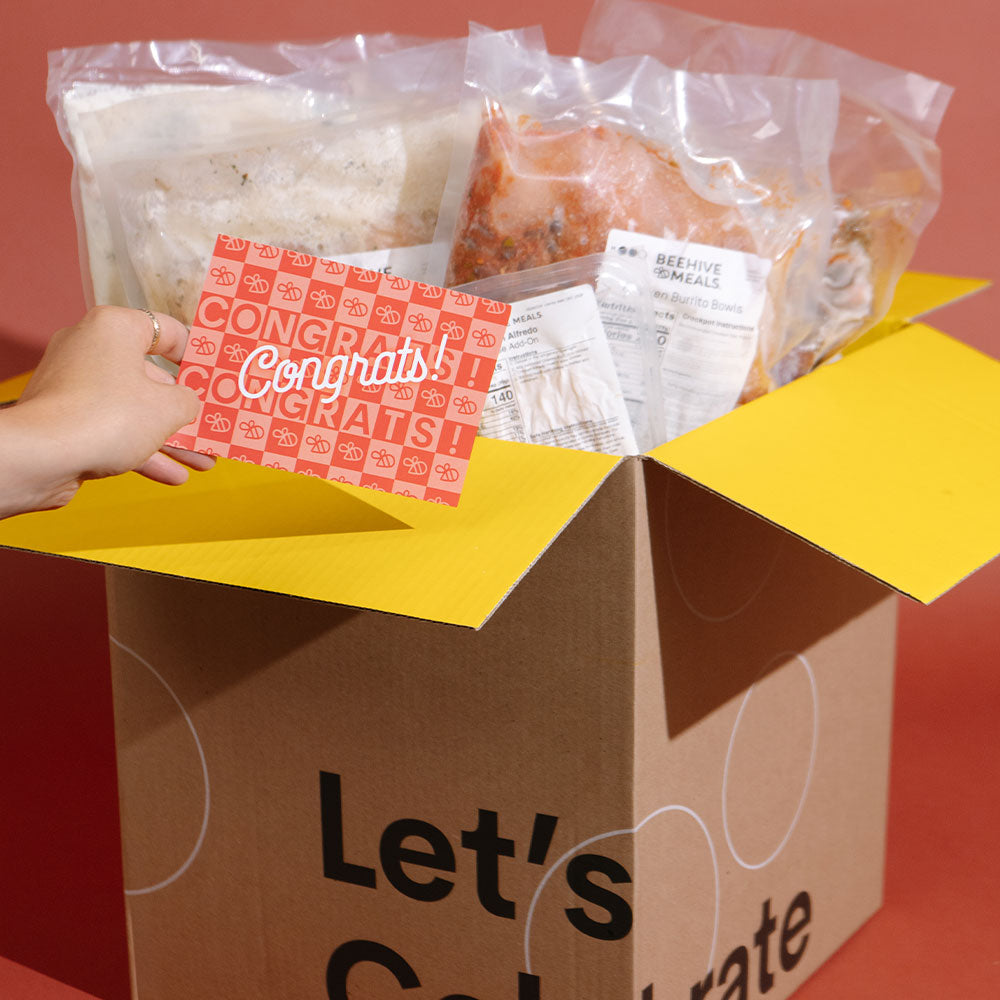 A cardboard gift box with yellow flaps open to reveal packaged food items and a red-and-white congratulatory card being held by a visible hand. The box front reads 'Let's Celebrate.