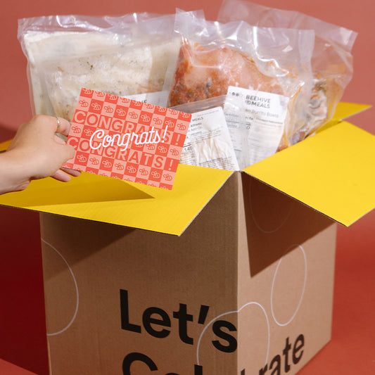 A cardboard gift box with yellow flaps open to reveal packaged food items and a red-and-white congratulatory card being held by a visible hand. The box front reads 'Let's Celebrate.