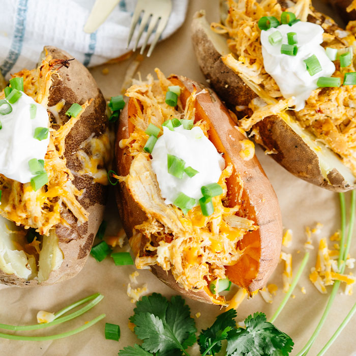 Baked potatoes stuffed with Beehive Meals Creamy Buffalo Chicken, topped with cheese, sour cream and green onions on a wooden surface.