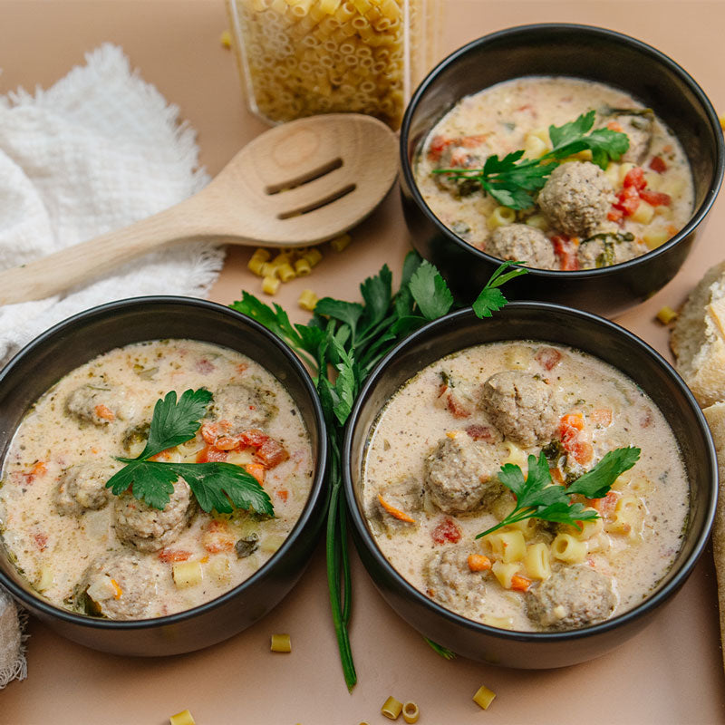 Three bowls of meatball soup with pasta and vegetables on a wooden surface.