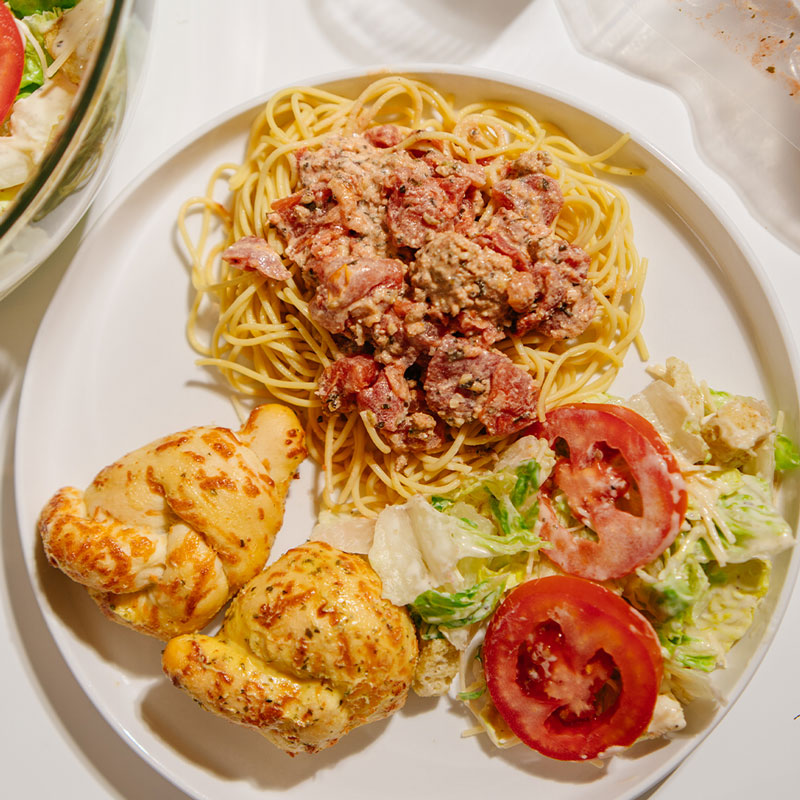 A top-down view of a white plate containing spaghetti topped with Beehive Meals Creamy Italian Sausage sauce, two garlic knots, and a side salad with tomato slices, set on a white table.