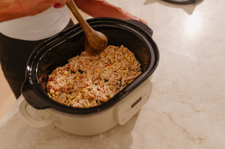 A person using a wooden spoon to stir a Beehive Meals meal inside a cream and black slow cooker on a light marble countertop.