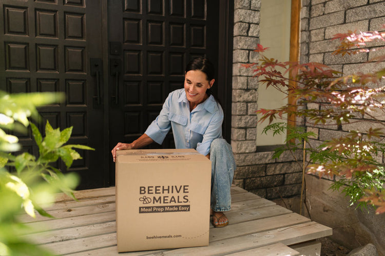 A woman sits on a wooden porch step holding a large Beehive Meals box, set against a background of a dark wooden door and stone wall.