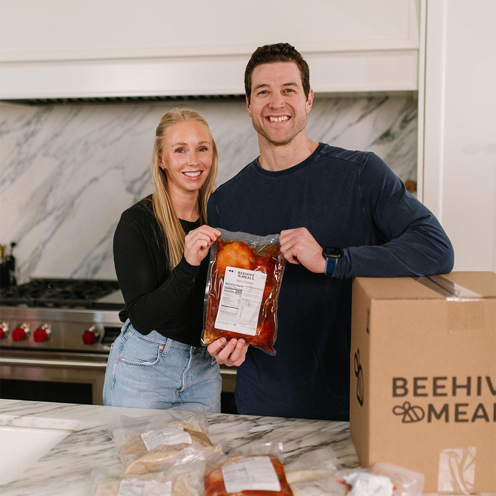 Two people holding a package of meat in a kitchen with a box labeled 'Beehive Meats' on a marble countertop.