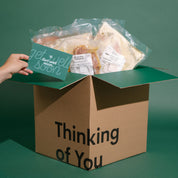 A cardboard gift box with the words 'Thinking of You' printed on the front, filled with packaged sandwiches and a teal card that says 'get well soon.' A hand is holding the card.
