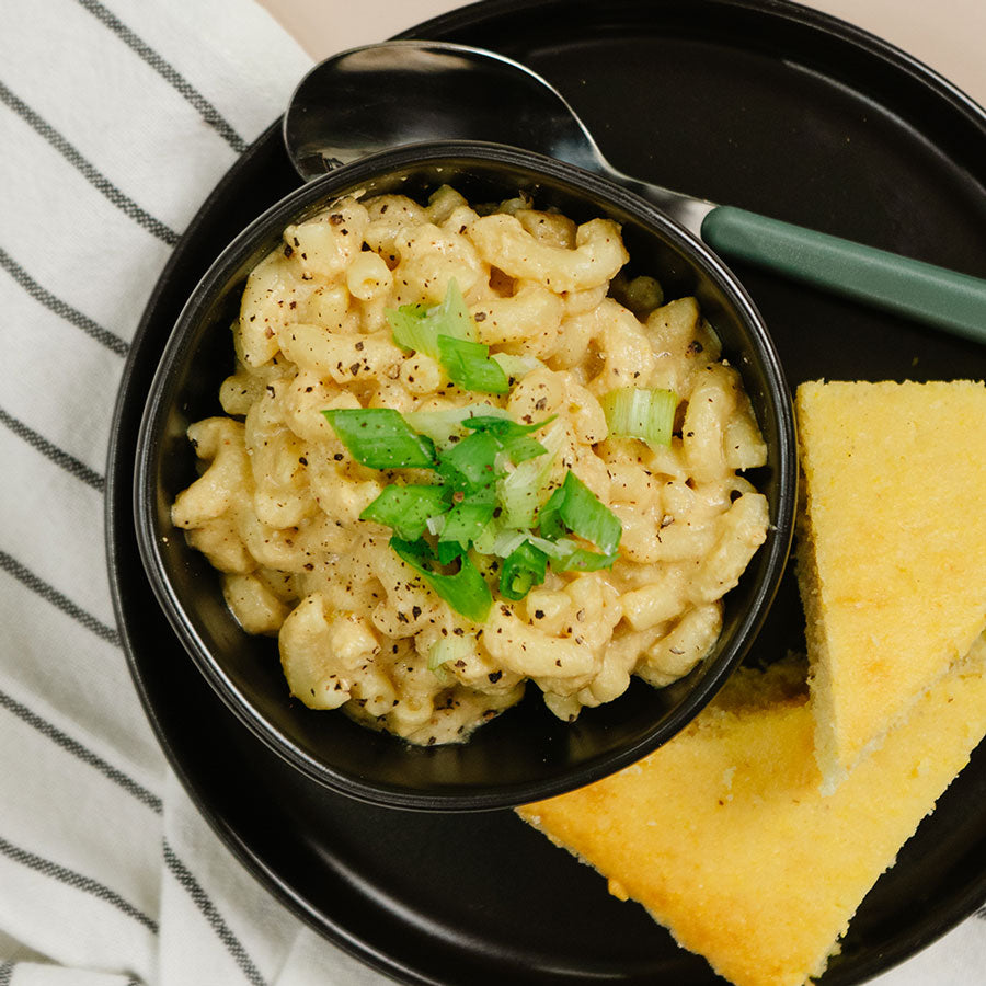 A bowl of creamy macaroni pasta topped with chopped green onions, served on a black plate with a spoon and slices of cornbread, next to a striped cloth napkin.
