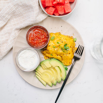 Plate with hash brown casserole, avocado slices, salsa, sour cream, and watermelon on a white surface.
