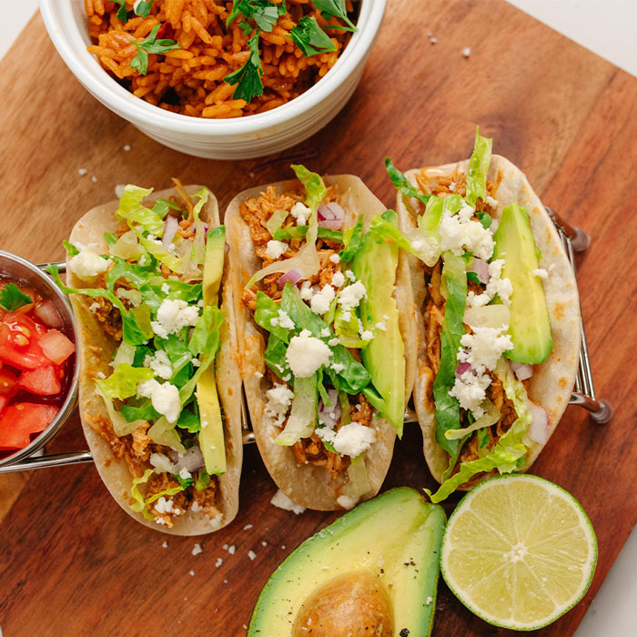 Three tacos with avocado and lime on a wooden board with side dishes.