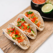 Three tacos with toppings on a wooden board with a lime and salsa in the background.