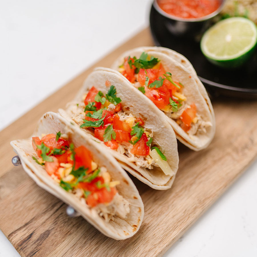 Three tacos with toppings on a wooden board with a lime and salsa in the background.