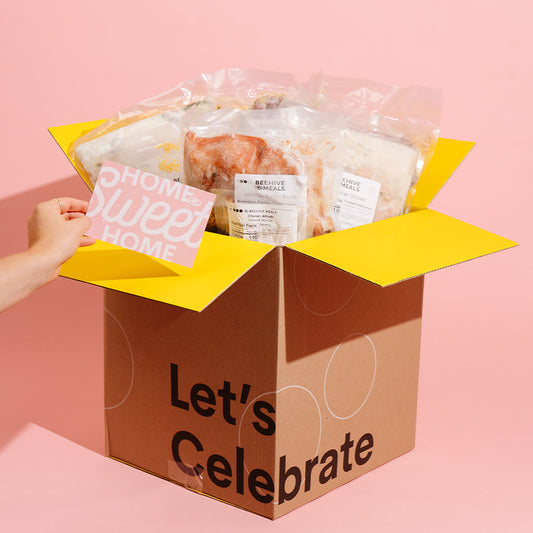 A brown cardboard gift box with yellow flaps and printed text reading 'Let's Celebrate' on the front, filled with packaged food items. A hand on the left holds a pink card that says 'Home Sweet Home.