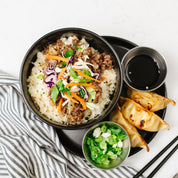 Beehive Meals Korean Egg Roll Bowls over rice topped with carrots and cabbage on a black plate with potstickers on the side