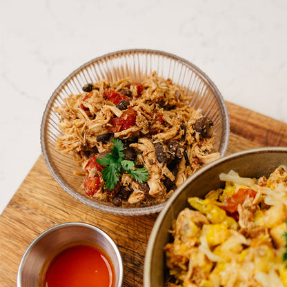 Two bowls of food on a wooden board with a small bowl of red sauce.