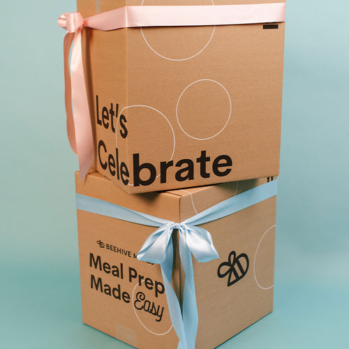 Two cardboard boxes with blue and pink ribbons and text 'lets celebrate' and 'meal prep made easy' on a light blue background