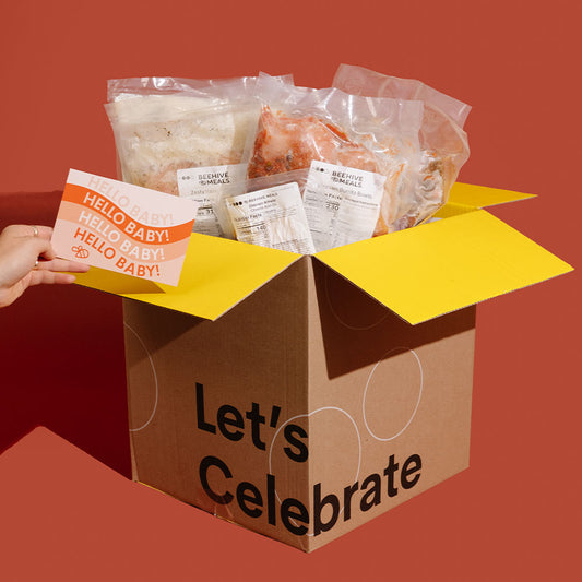 A brown cardboard celebration box with yellow flaps and the words 'Let's Celebrate' printed on the front, filled with packaged food items and a small printed card held by a visible hand. The background is a solid warm red-orange studio backdrop.
