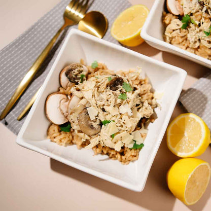 White square bowl filled with Beehive Meals Parmesan Herb Chicken over rice on a beige surface with lemons and gold cutlery.