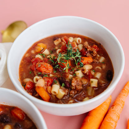 Bowl of Beehive Meals Pasta Fagioli soup with carrots on a pink background