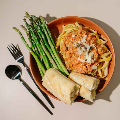 Dinner plate with Beehive Meals Sausage Marinara served over fehttuchini noodles with a side of asparagus and bread on a beige background with a black fork and spoon