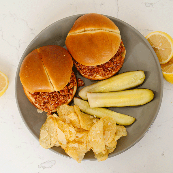 Two hamburger buns filled with Beehive Meals Sloppy Joes with chips and pickle spears on the side all on a grey plate on a marble background.