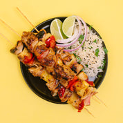 Grilled chicken and pineapple skewers served on a black plate with white rice, sliced red onion, lime wedges, and black sesame seeds, set against a solid yellow background.