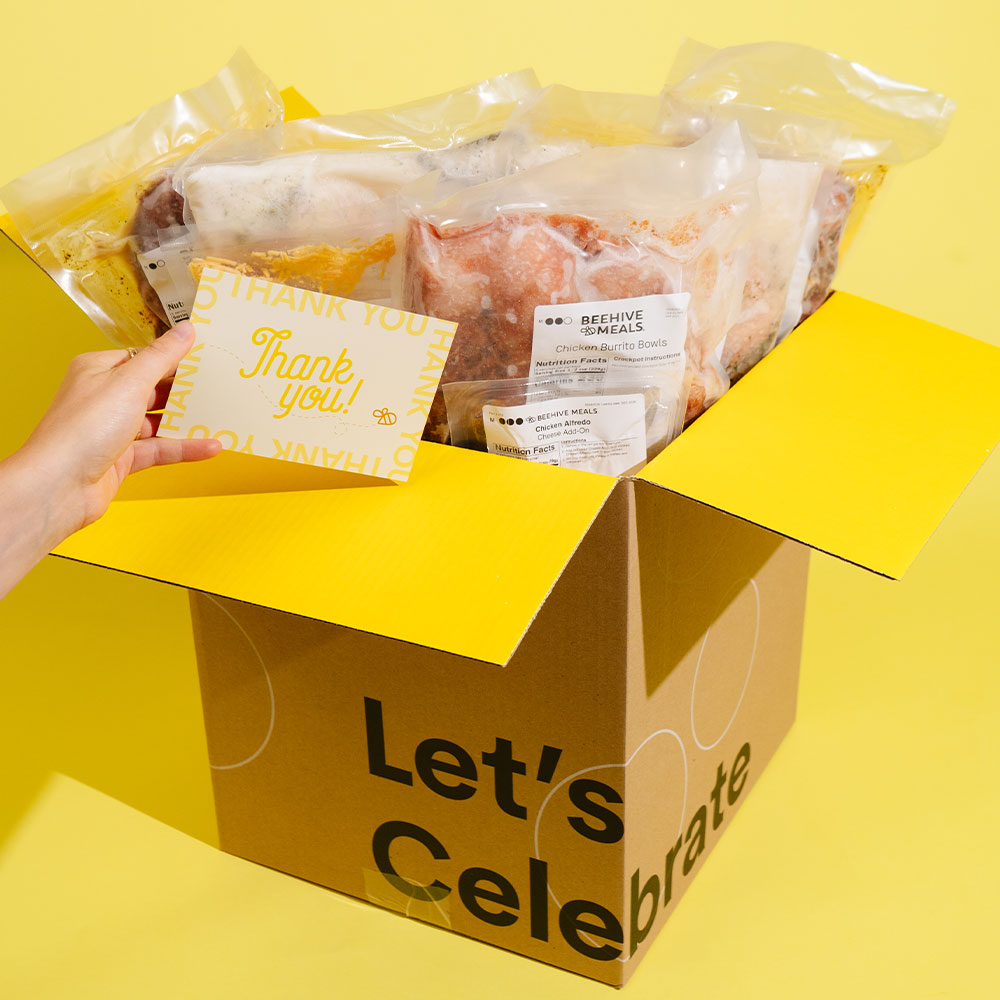 A yellow and brown cardboard celebration box labeled 'Let's Celebrate' filled with packaged food items, with a hand holding a yellow 'Thank you' card inside. The background is a solid yellow studio backdrop.
