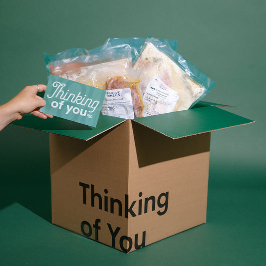 A cardboard gift box printed with the words 'Thinking of You' sits open on a dark green background. A hand on the left holds a matching teal card inside the box, which is filled with packaged food items.