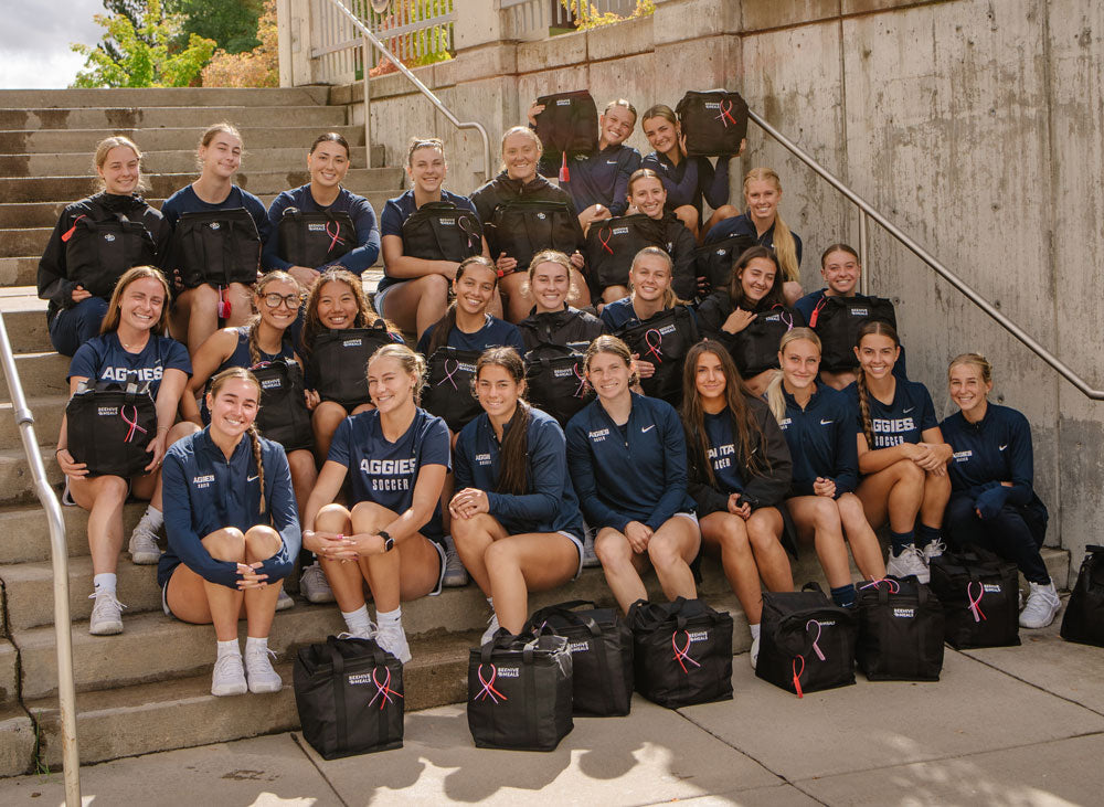 A group of female Utah State University athletes posing together on outdoor concrete stairs, wearing matching navy blue athletic jackets and shorts, with black Beehive Meals cooler bags in front of them. 