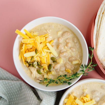 Bowl of Beehive Meals White Bean Chicken Chili. It's a creamy soup with cheese and herbs on a pink background