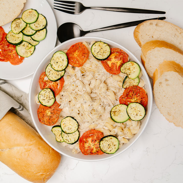 White bowl with noodles and Beehive Meals Zesty Italian Chicken with zucchini and tomatoes on a white table with bread and cutlery.