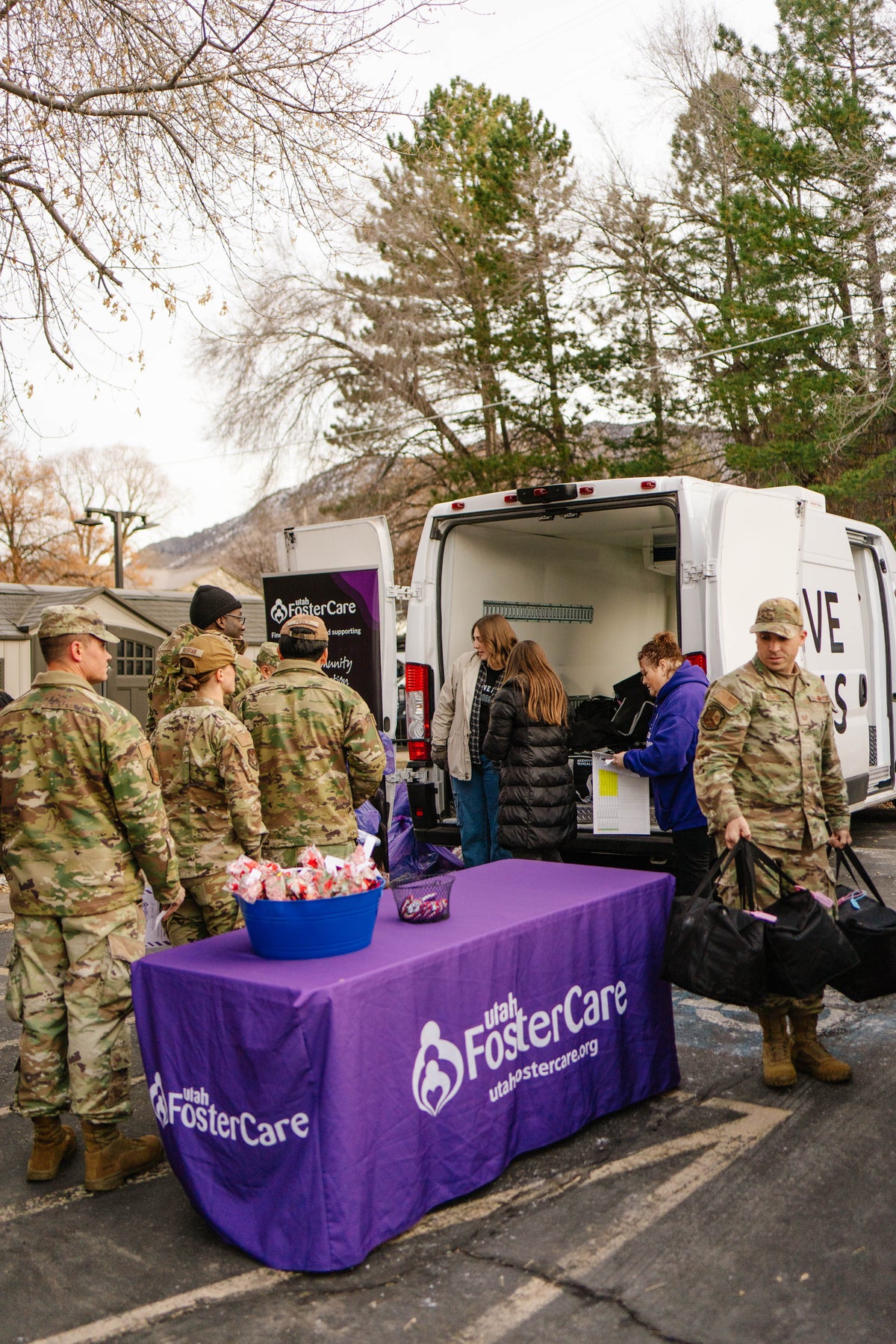 A group of people in military uniforms gathered around a purple table covered with a FosterCare-branded cloth, standing beside an Beehive Meals van. 