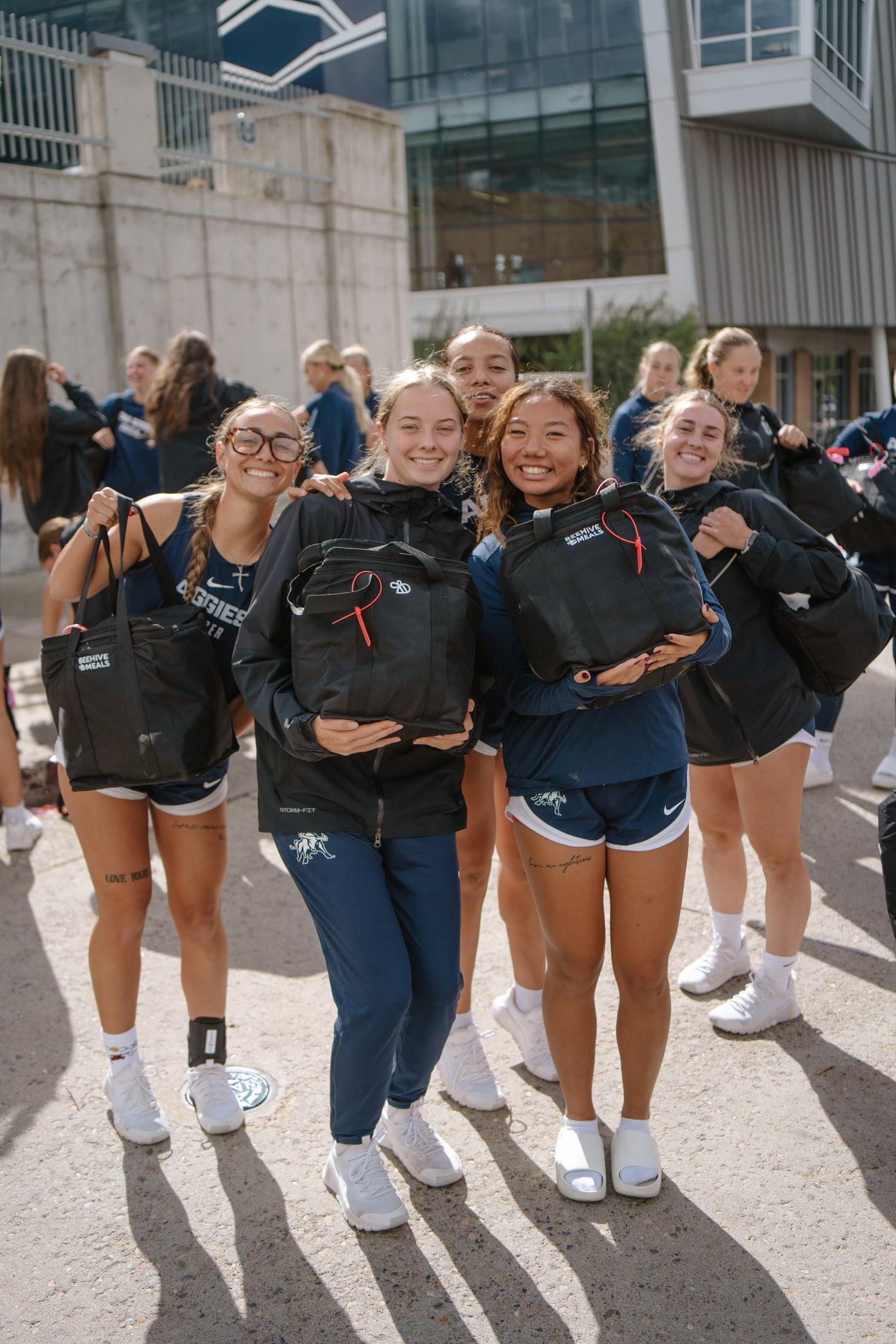 A group of young female athletes posing outdoors on a sunny day, each holding a Beehive Meals bag. 