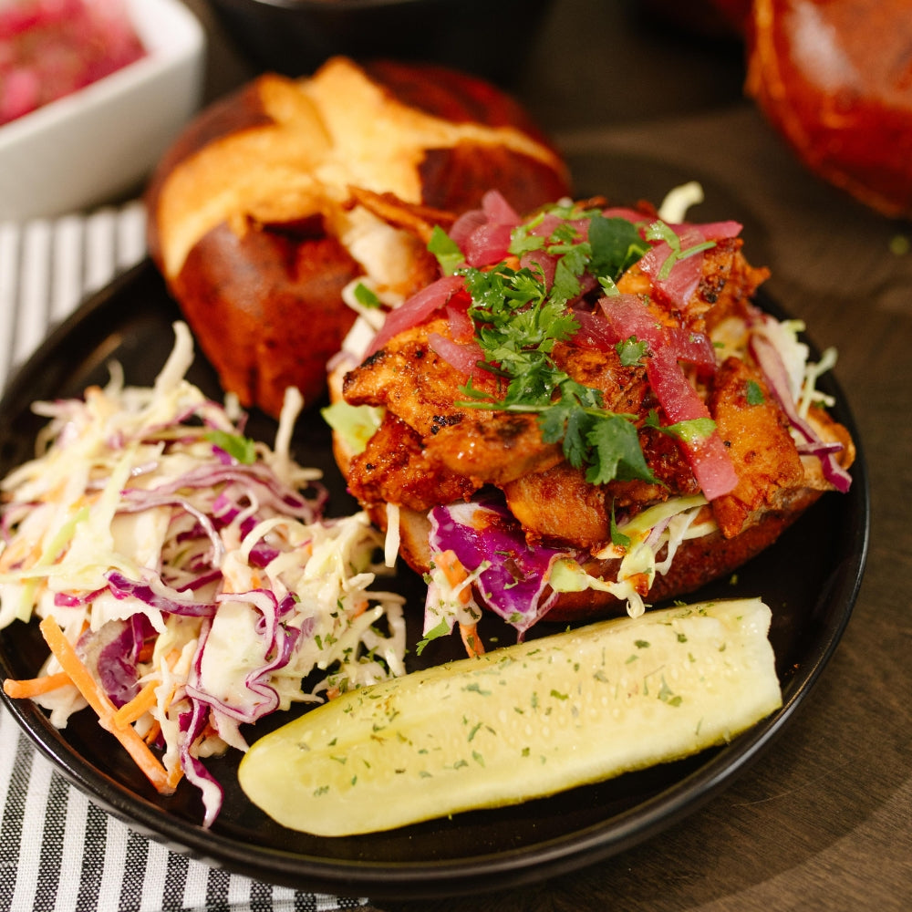 Plated BBQ Chicken Sandwich with pickled onions, cilantro, coleslaw, and a pickle on the side.