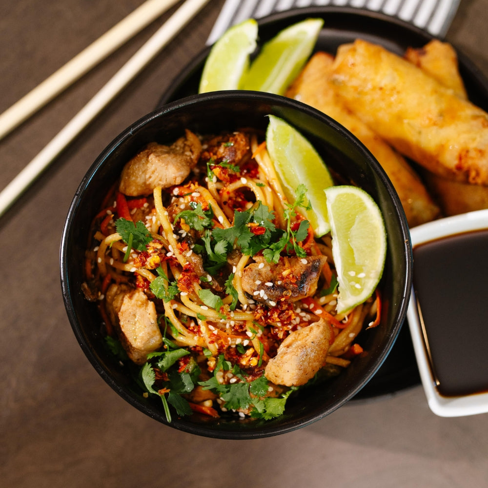 Bowl of Beehive Meals Chicken Lo Mein with noodles, lime wedges, garnished with herbs, on a wooden table.
