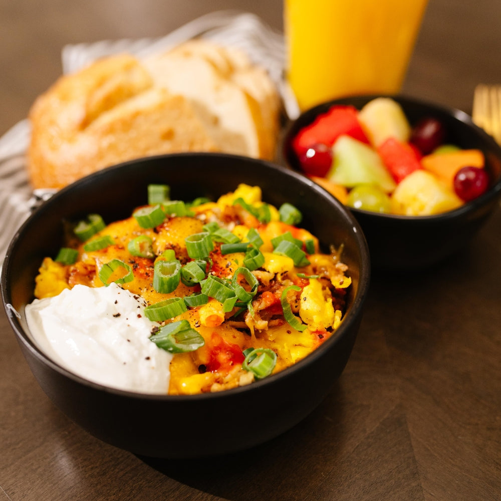 Bowl of Beehive Meals Sausage Griddle Hash Browns with green onions, sour cream, and a side of fruit and bread on a wooden table.