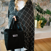 Person wrapped in a black and white grid-patterned blanket holding a black tote bag with 'Beehive Meals' logo.